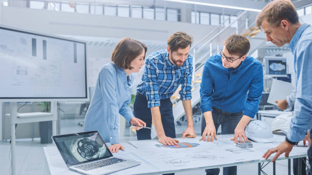 OPD DESIGN | What Does an Industrial Design Company Actually Do? In the Industrial Engineering Facility: Diverse Group of Engineers and Technicians on a Meeting Gather Around Table with Engine Design Technical Drafts, Have Discussion, Analyse Technology